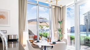 A modern dining area with a large window, a view of a pool, and a dining table set with plates and glasses. - LUX The District One Lagoon View Suite (Dubai)
