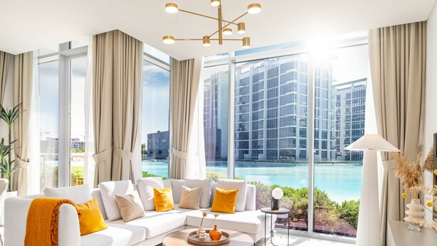 A modern living room with a white sofa, yellow cushions, and a view of a building and a pool. - LUX The District One Lagoon View Suite (Dubai)
