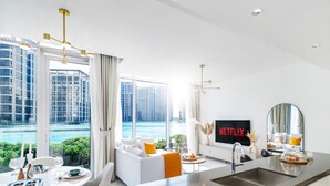 A modern living room with a large TV displaying 'NETFLIX', a dining area with a view of the water, and a large window with curtains. - LUX The District One Lagoon View Suite (Dubai)
