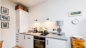 A modern kitchen with white cabinets, a built-in oven, a gas stove, and a sink. There are framed pictures on the wall, a clock, and a small wooden table with a bowl. - Delightful 1-bed, Pimlico (London)