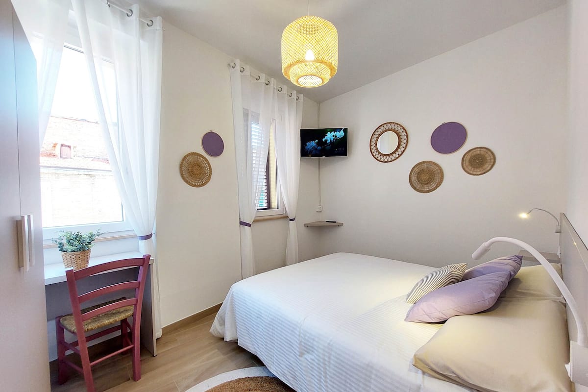 A bedroom with a bed, a TV mounted on the wall, a window with curtains, a chair, and a small shelf.