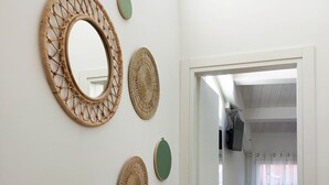Un pasillo con una pared blanca decorada con arte de pared tejido y un espejo. Una puerta da a una habitación con una silla verde y una pequeña mesa con una planta.