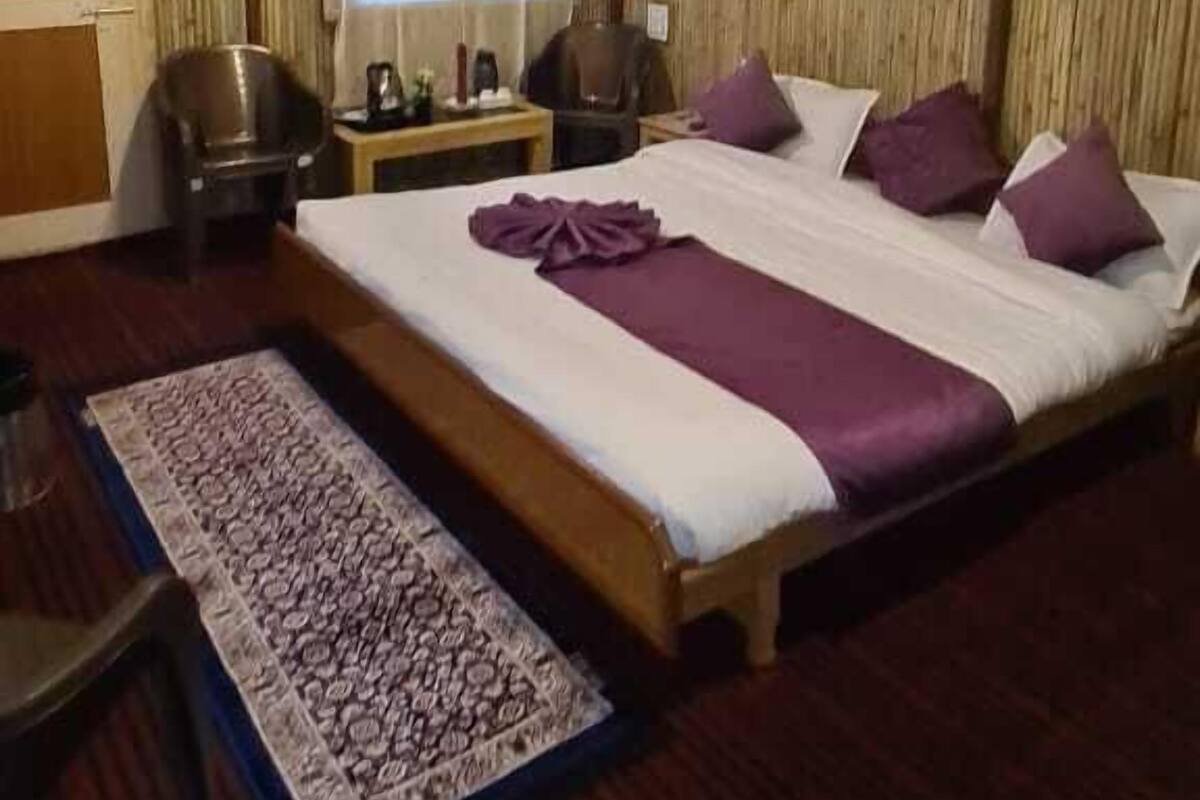 A bedroom with a wooden ceiling, a large bed with purple bedding, a small table, a chair, and a window with curtains.