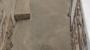 Un patio estrecho con un muro de piedra, un pequeño jardín con flores y una tapa de alcantarilla.