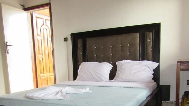 Una cama con cabecera oscura y ropa de cama blanca, una puerta de madera al fondo y un enchufe visible en la pared.