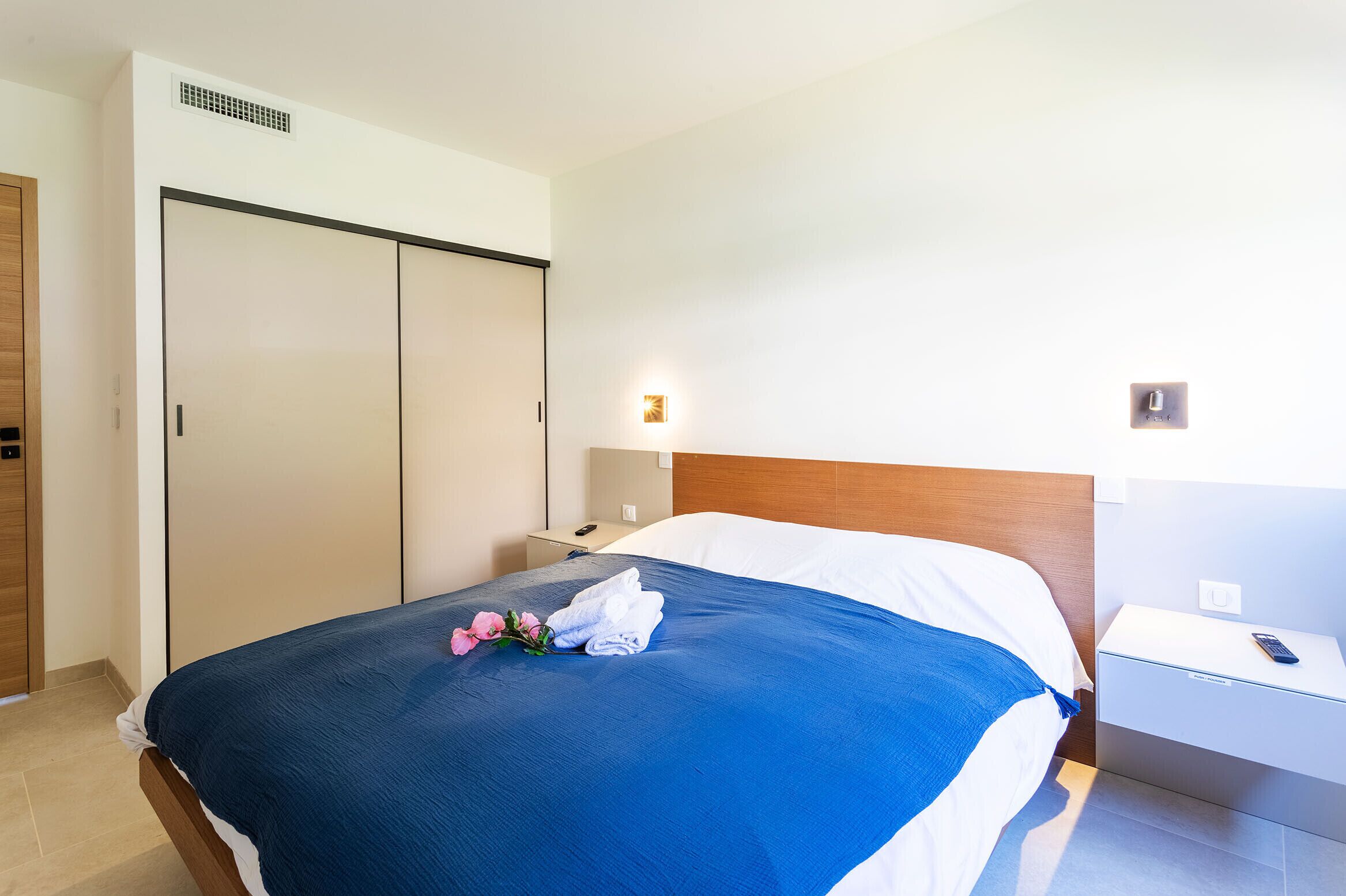 Une chambre d’hôtel comprenant un lit, une table de chevet, une lampe de chevet, une armoire et un luminaire fixé au mur.