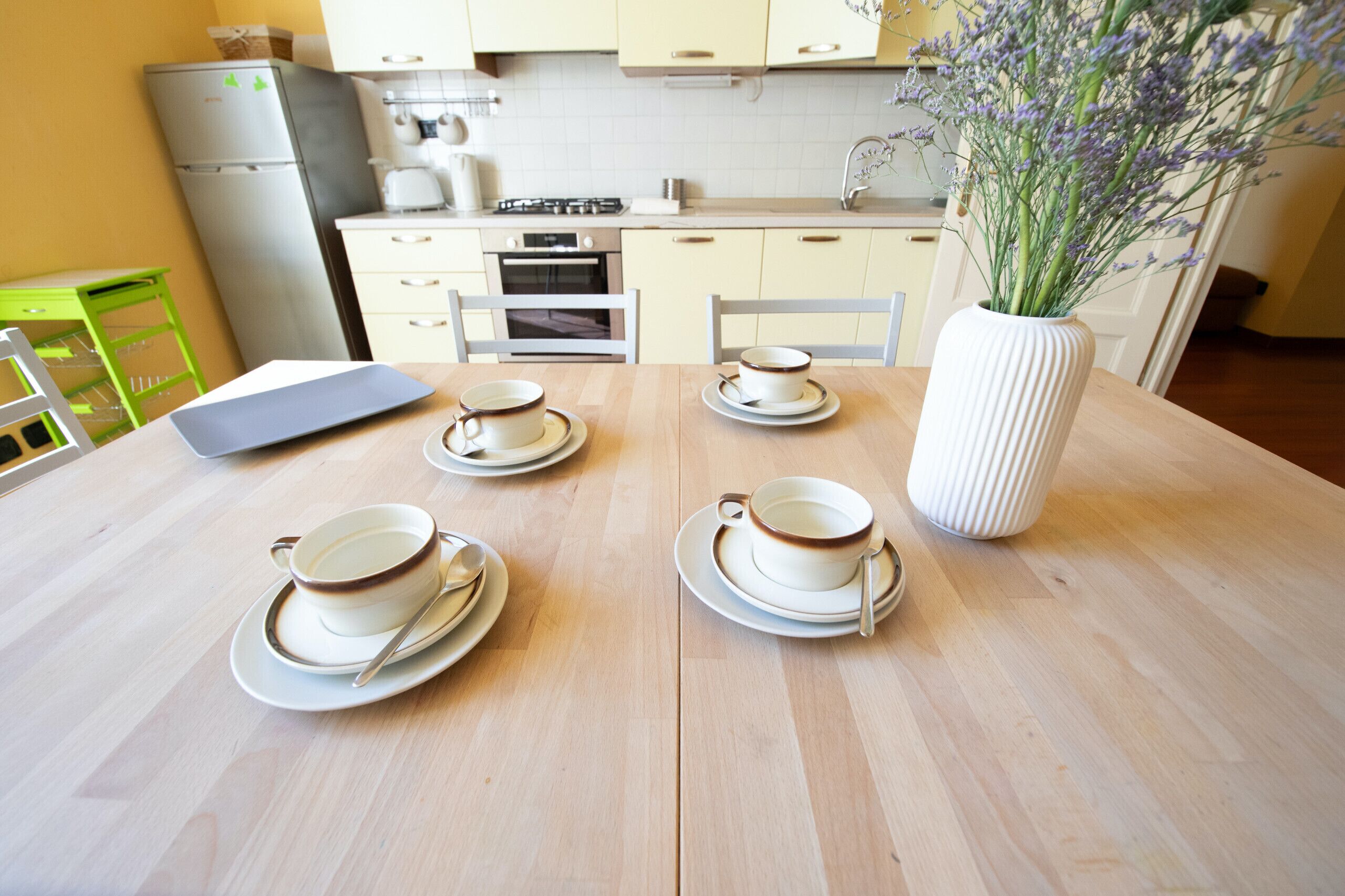 A dining table set with four cups and saucers, a vase with flowers, and a laptop.