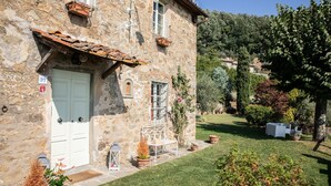 A stone building with a white door, a small porch with a white bench, and a well-maintained lawn. - Villa Iris with Swimming Pool (Capannori)