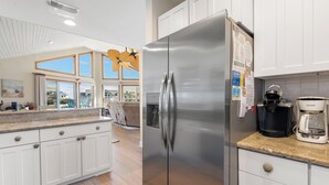 Eine moderne Küche mit weißen Schränken, einem Edelstahl-Kühlschrank, einer Kaffeemaschine und Blick durch große Fenster nach draußen.