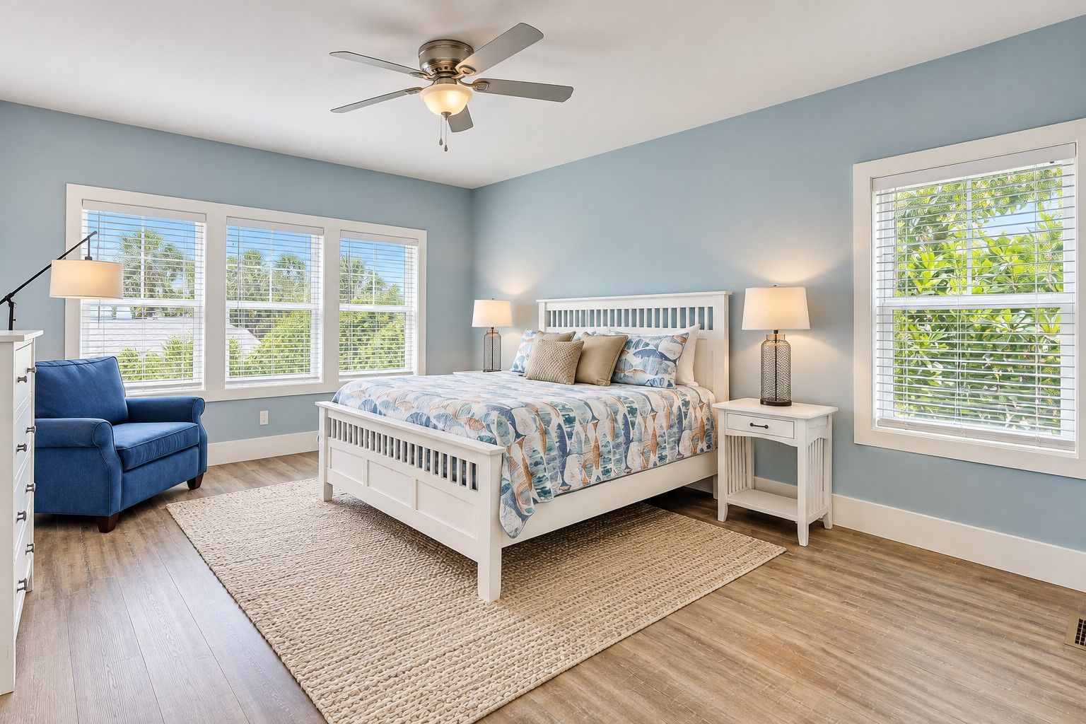 A bedroom with a bed, two bedside tables, a chair, a ceiling fan, and large windows with blinds.