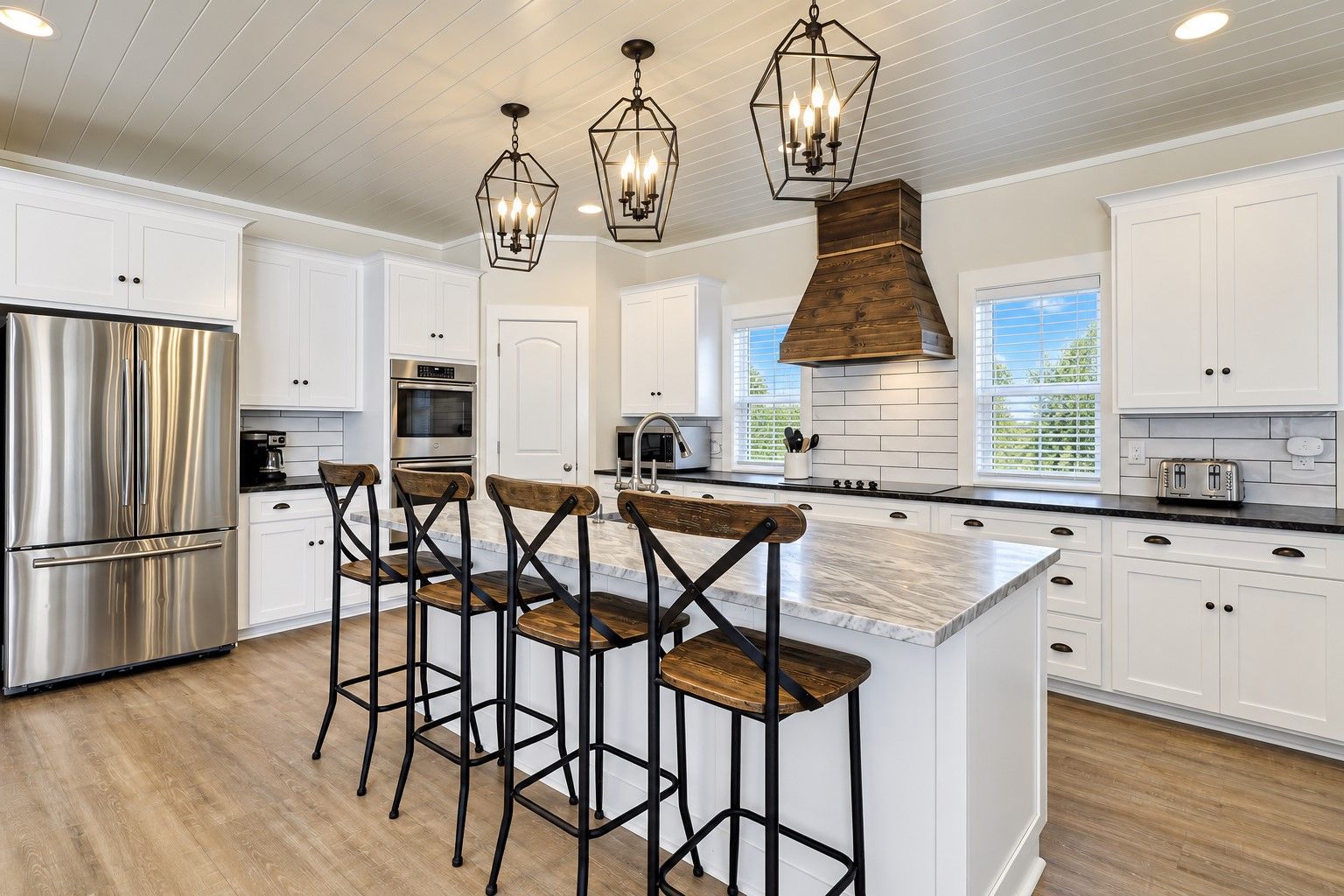 A modern kitchen with white cabinetry, a stainless steel refrigerator, a wooden island with bar stools, and pendant lighting.