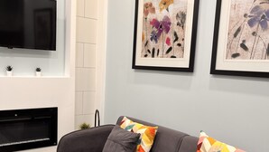 A modern living room with a grey sofa, two framed floral paintings, a flat-screen TV, a fireplace, and a wooden coffee table. - One bedroom apartment/suite  (Whitby)