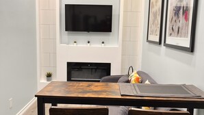 A modern living room with a flat-screen TV mounted on a white brick wall, a wooden dining table with bar stools, and a fireplace below the TV. - One bedroom apartment/suite  (Whitby)
