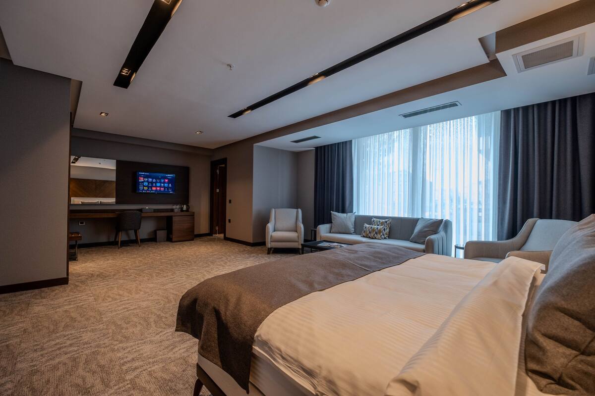 A modern hotel room with a large bed, a desk with a computer, a sofa, and a TV.