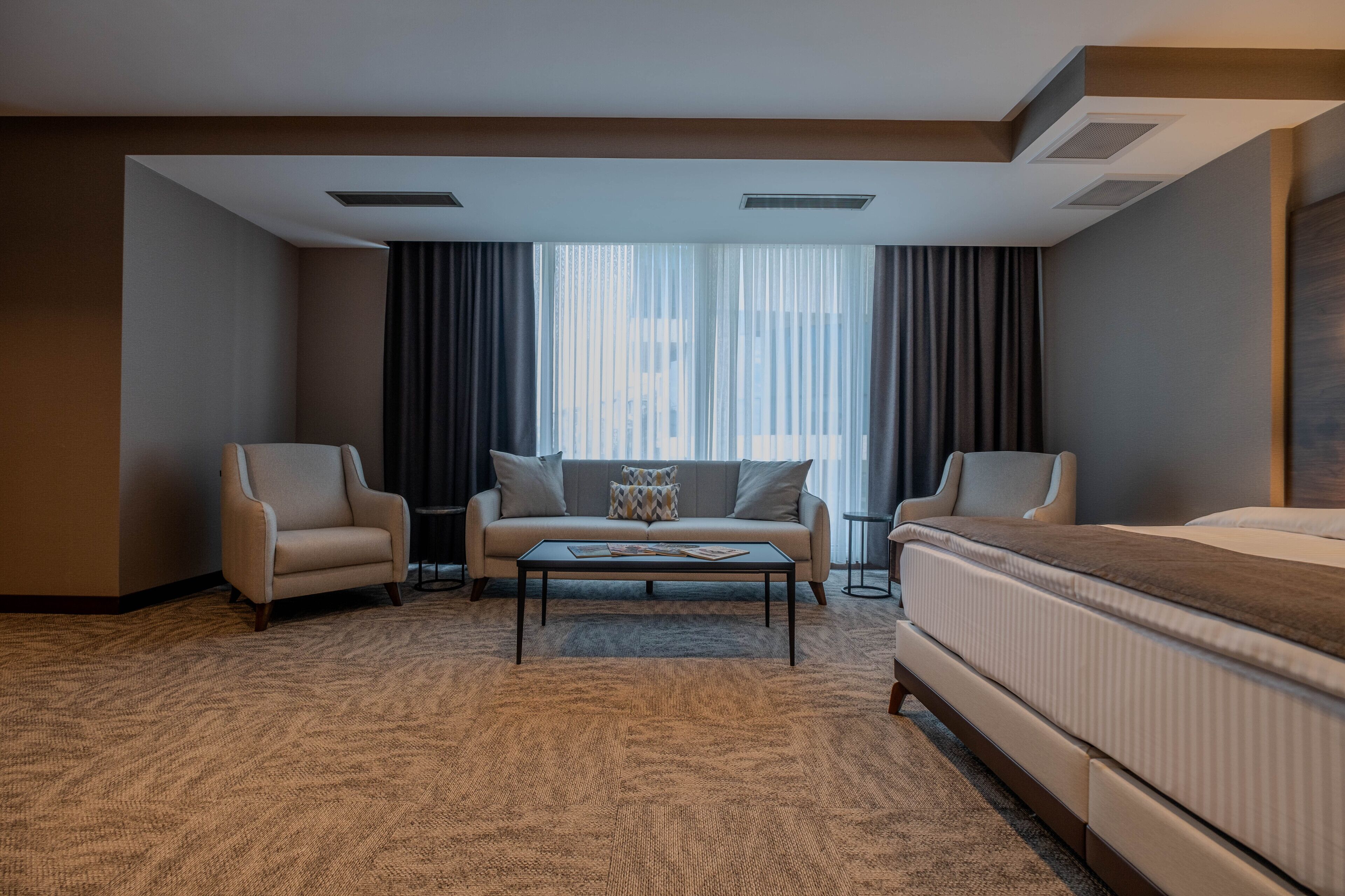 Habitación de hotel con cama, dos sillones, sofá, una mesa de centro y un ventanal con cortinas.