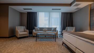 Habitación de hotel con cama, dos sillones, sofá, una mesa de centro y un ventanal con cortinas.