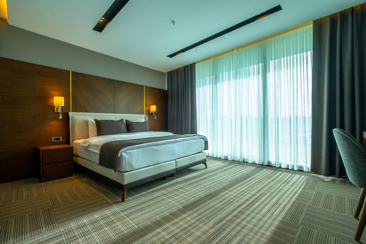 A modern hotel room with a large bed, two bedside lamps, a nightstand, a chair, and a large window with curtains.