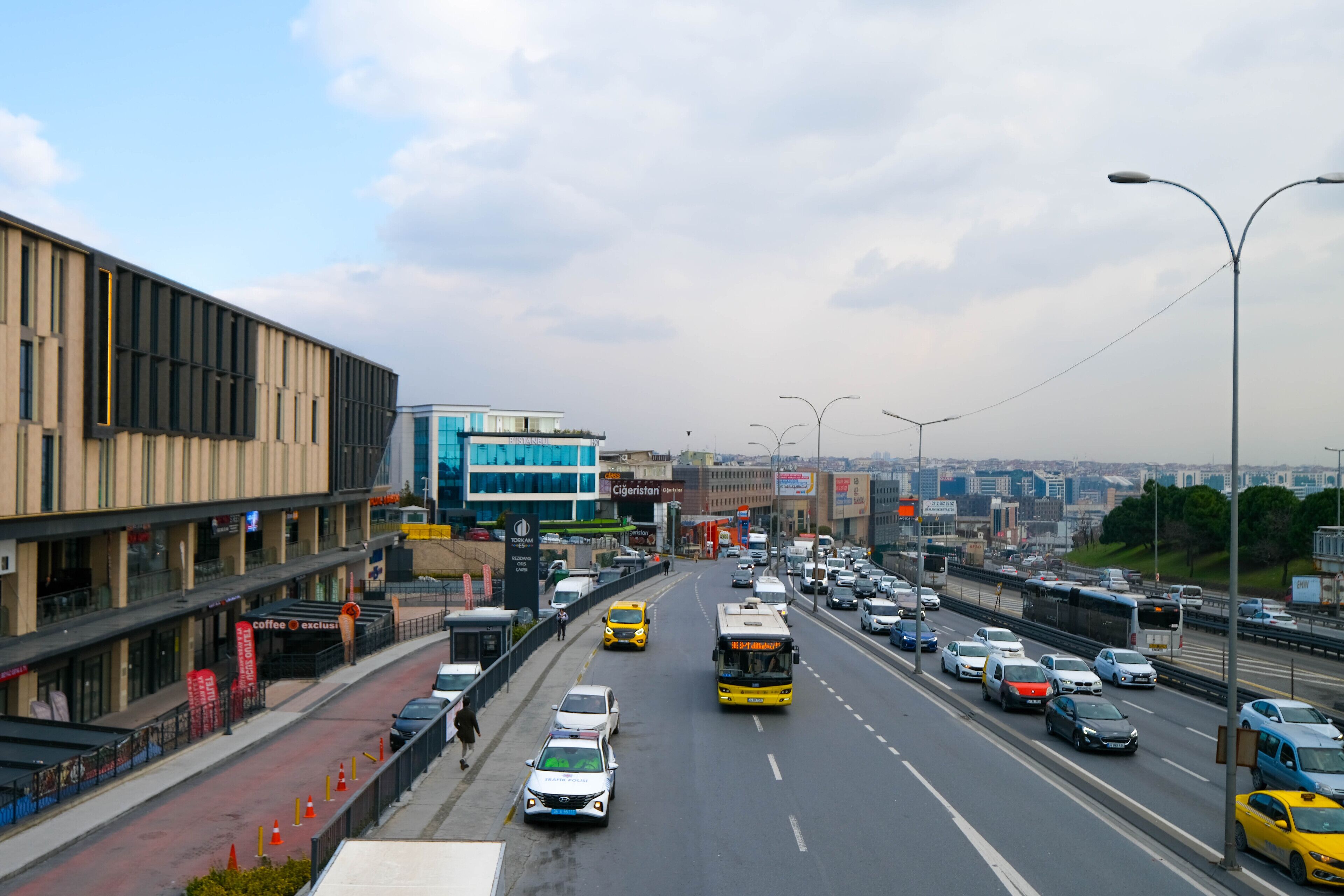A busy urban street with multiple lanes of traffic, including cars, buses, and taxis. There are modern buildings with large windows and signage on the left side.