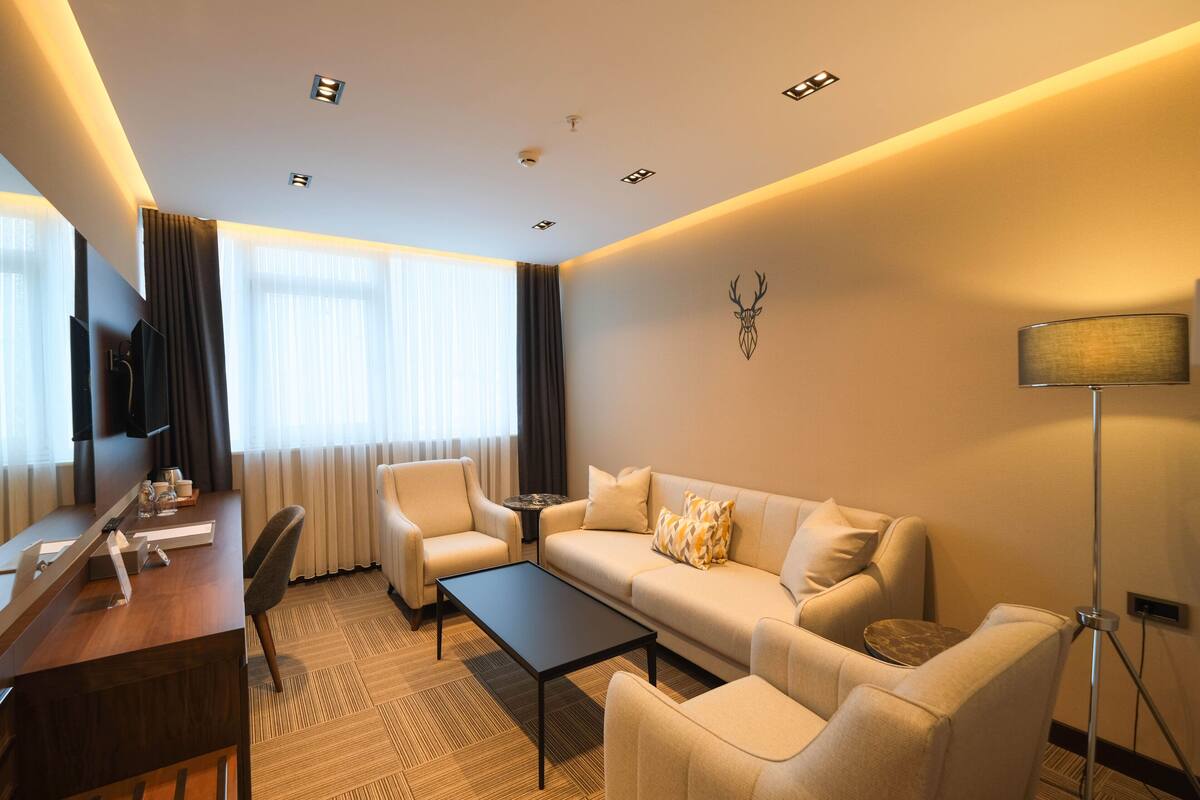 A modern hotel room with a sofa, armchairs, a coffee table, a desk with a flat-screen TV, and a decorative wall piece.