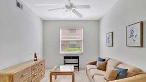 A living room with a ceiling fan, a window with blinds, a wooden cabinet, a glass-top coffee table, and a beige sofa with cushions. - Cypress Woods for Seasonal or Summer Stays W Golf (Naples)