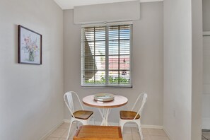 Un petit coin repas avec une table ronde et deux chaises, une fenêtre avec des stores et un tableau encadré au mur.