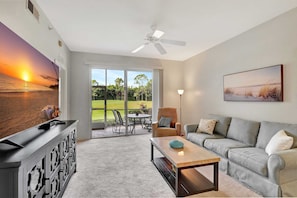 A living room with a large TV, a ceiling fan, a sofa, a coffee table, and a view of a garden through the glass door.