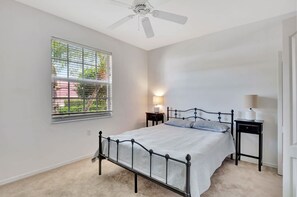 A bedroom with a bed, a ceiling fan, a window with blinds, and a nightstand with a lamp.