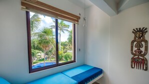 A room with a window overlooking a pool and palm trees, a blue bench, and a wooden sculpture on the wall. - Family getaway in this charming townhouse just steps away from Yal Ku Laggon! (Akumal)