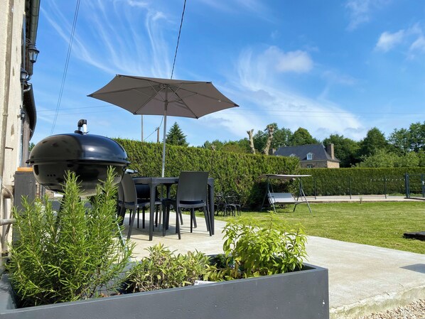 Um pátio externo com churrasqueira, mesa de jantar, cadeiras e um guarda-sol.