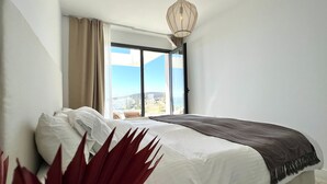 Un dormitorio con una cama grande, vistas de la ciudad a través de un ventanal corredera y una lámpara colgante.