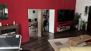 Ein modernes Wohnzimmer mit roter Akzentwand, einem Flachbildfernseher, einem Holztisch und einem Essbereich, der durch einen offenen Durchgang sichtbar ist.