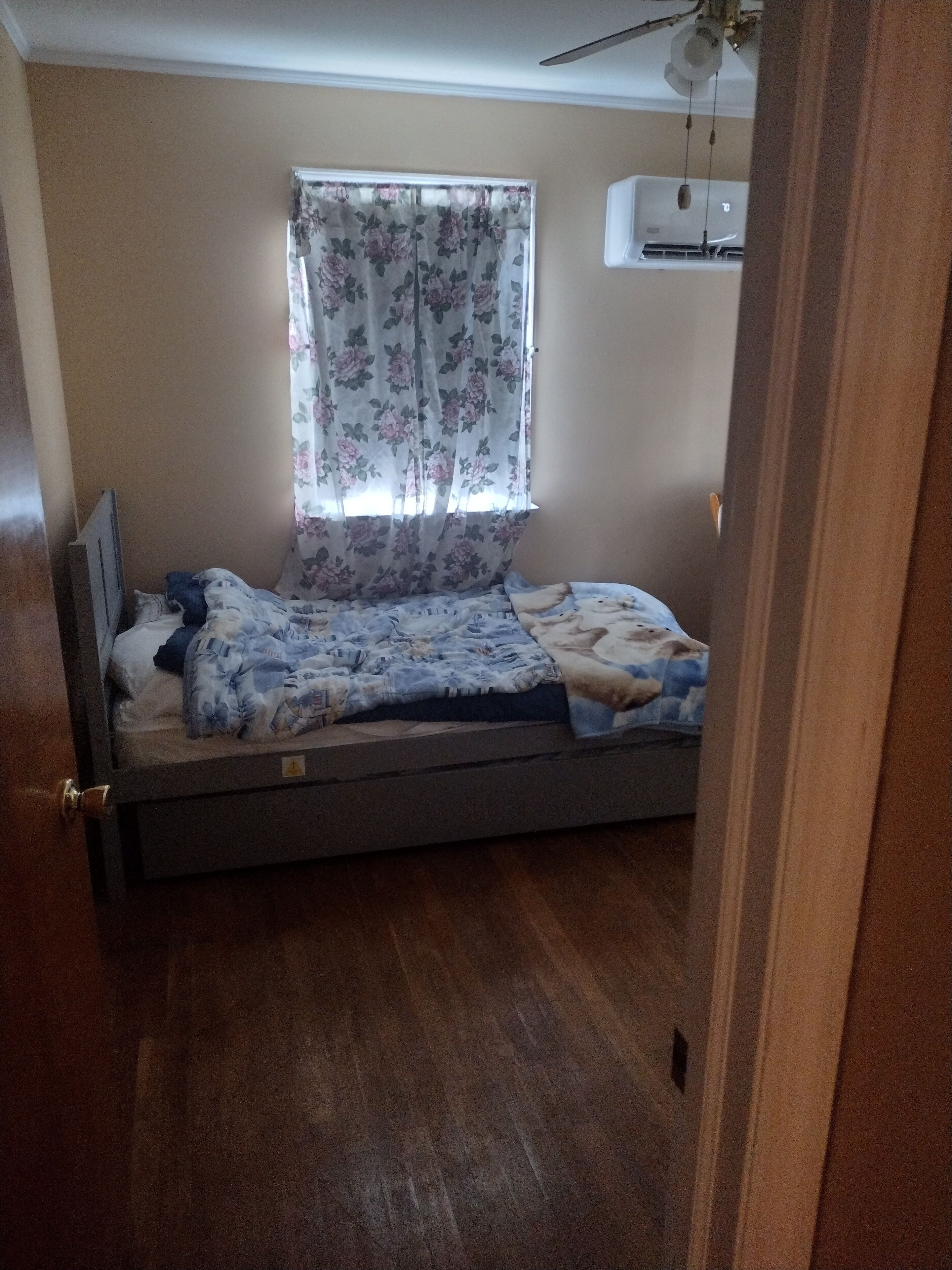 A single bed with a floral-patterned bedspread, a window with floral curtains, and an air conditioning unit mounted on the wall.