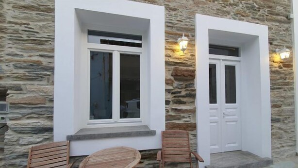 A stone building with two white doors, each with a window and a light fixture above. There are two wooden chairs and a small table outside. - Cozy Loft in Chora of Andros 1 min from the main beach! (Andros)