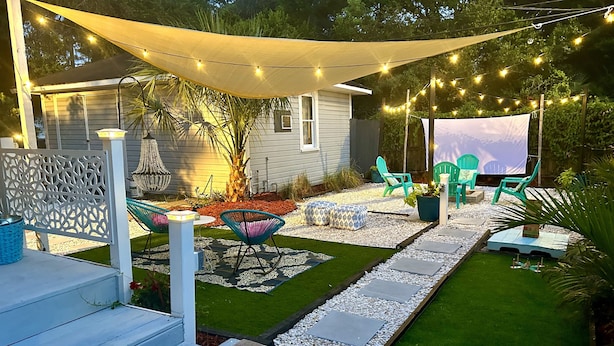 Un patio trasero con mobiliario exterior, un pequeño jardín y luces colgantes.
