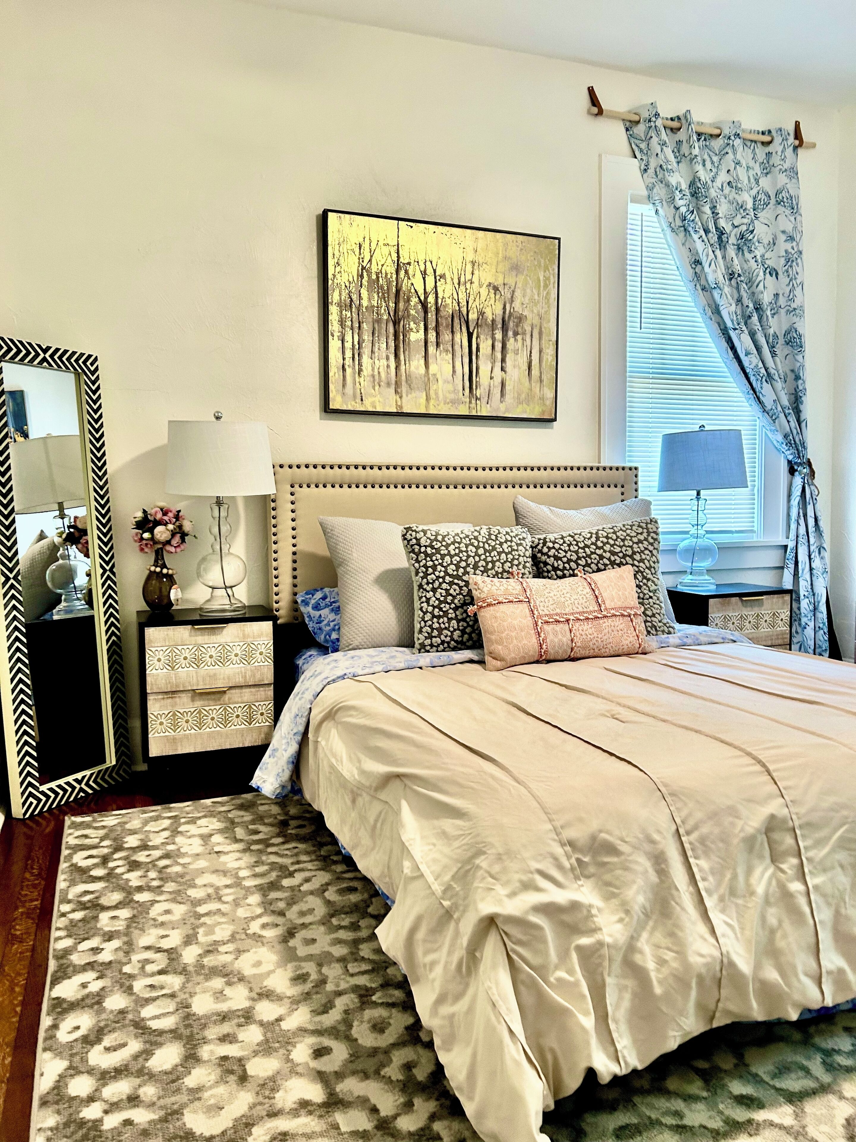 A neatly arranged bedroom with a bed, a bedside table with a lamp, a dresser, a mirror, and a window with curtains.