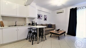 A modern kitchen with white cabinets, a dining area with a table and stools, a sofa, and a coffee table. - IQ Kleovoulou (Athens)