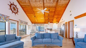 A spacious living area with a wooden ceiling, a large sofa, a coffee table, and a view of the kitchen and dining area.
