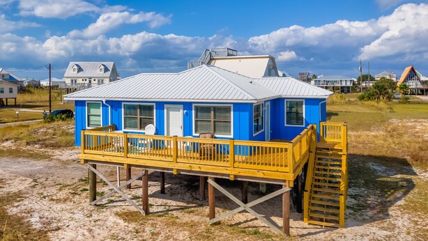 Ein blaues Haus mit einer Holzterrasse und einem Metalldach, das auf Stelzen über sandigem Grund steht.
