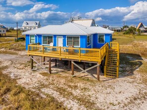 A blue house with a wooden deck and a metal roof, situated on stilts over sandy ground. - Boa Vista - 106 Ponchartrain 3 Bedroom Home by RedAwning (Dauphin Island)