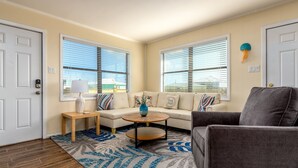 Ein Wohnzimmer mit Sofa, Sessel, Couchtisch und Teppich. Zwei Fenster mit Jalousien und eine Tür sind vorhanden.