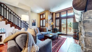 Un salon spacieux comprenant un escalier, un ensemble de canapés, une table basse et une grande fenêtre avec des stores.