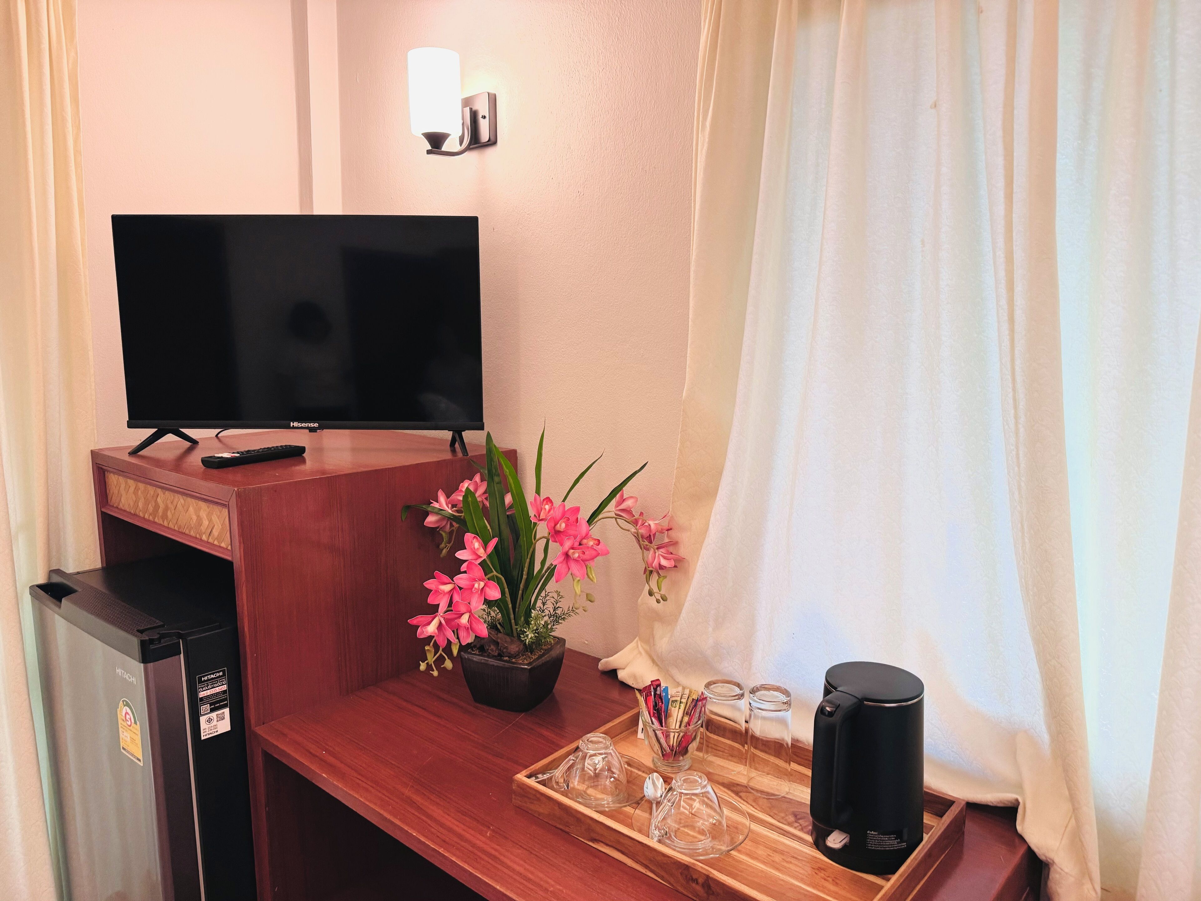 A hotel room with a flat-screen TV, a minibar, a wooden desk with a tray of glasses and a coffee maker, and a potted orchid plant.
