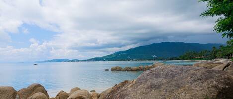 A coastal landscape with large rocks in the foreground, calm water, and distant mountains under a cloudy sky.