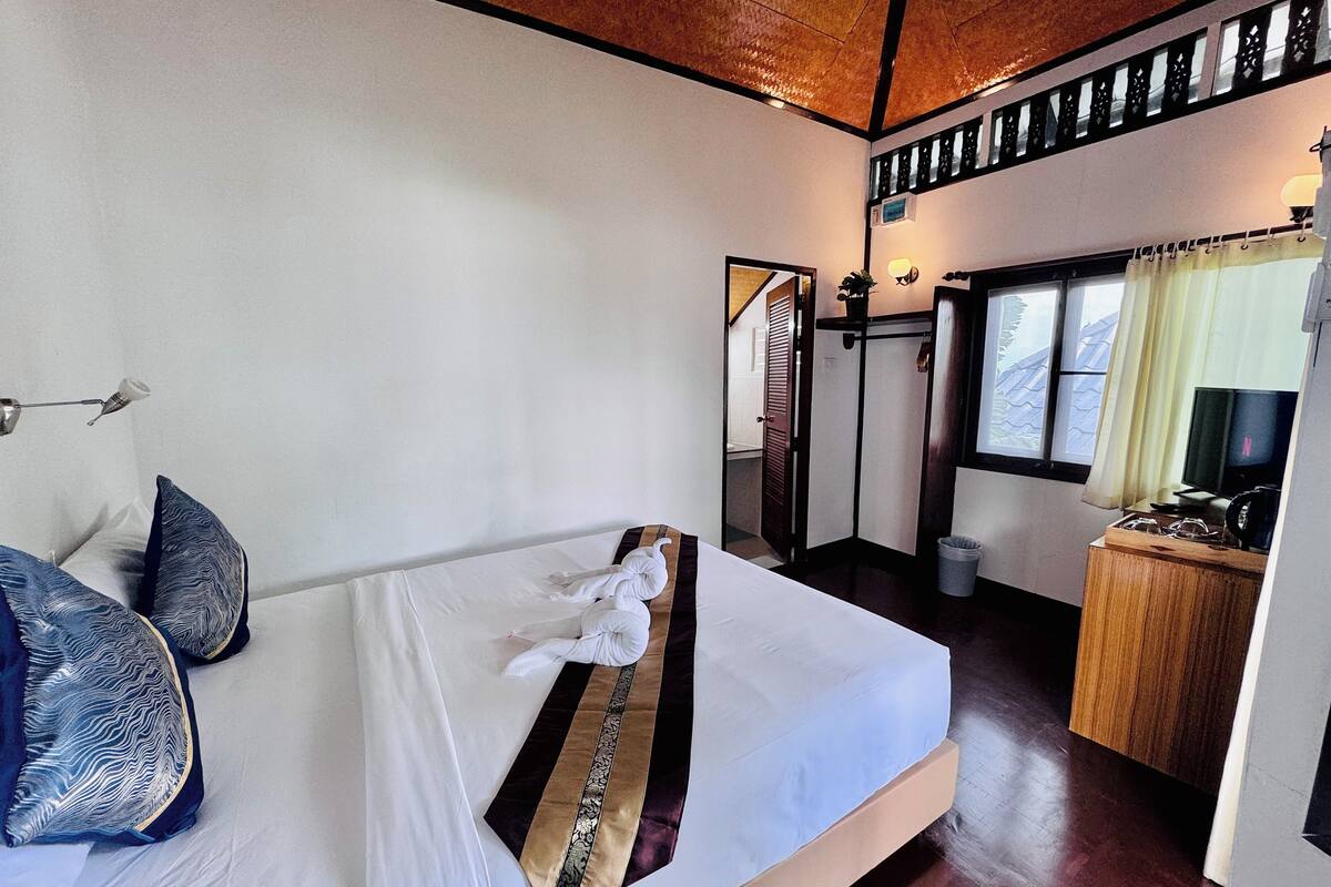 Quarto de hotel com cama, almofadas, escrivaninha de madeira, televisão e janela com cortinas.