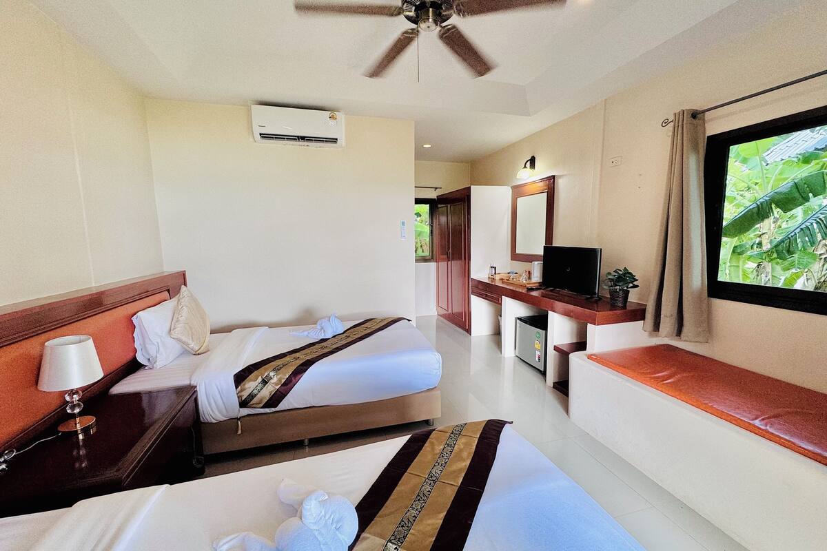 Quarto de hotel com duas camas, ventoinha no teto, ar condicionado, televisão, pequena escrivaninha, janela com vista para vegetação e banheiro com chuveiro e vaso sanitário.