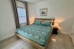 A bedroom with a bed, a nightstand, a lamp, a window with blinds, and a framed picture on the wall.