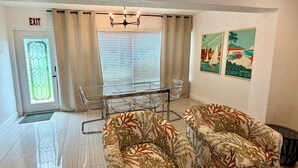 A room with a glass table, two patterned chairs, a window with blinds, a door with an exit sign, and two wall art pieces. - 3/3 Duplex for 6+Shared Heated Pool, Tiki Bar BBQ (Hollywood)
