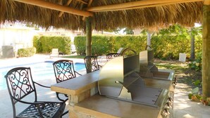An outdoor patio with a grill and seating area, a pool in the background, and a thatched roof structure. - 3/3 Duplex for 6+Shared Heated Pool, Tiki Bar BBQ (Hollywood)