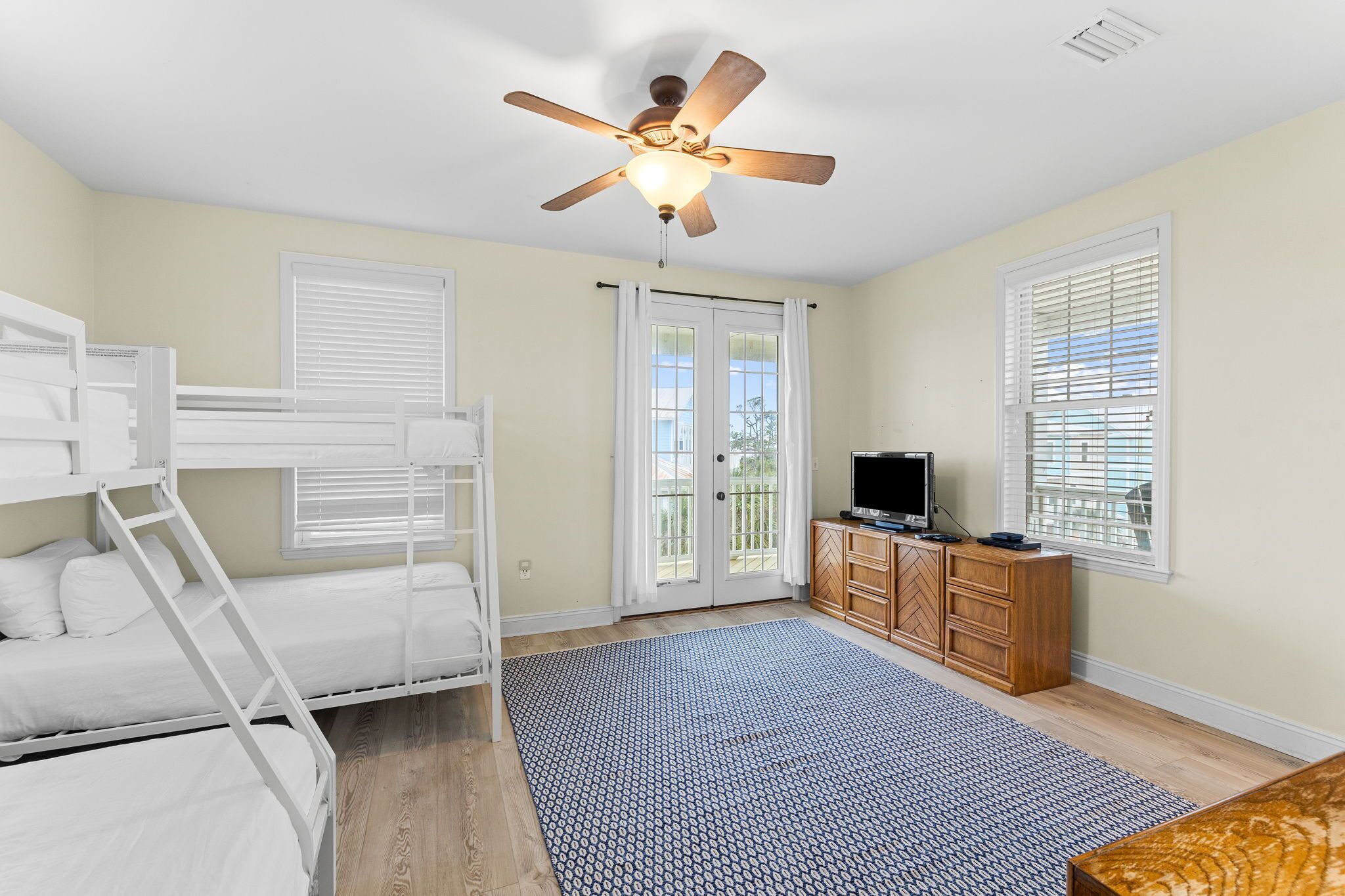 A room with a bunk bed, a ceiling fan, a television, a wooden cabinet, and a large window with blinds.