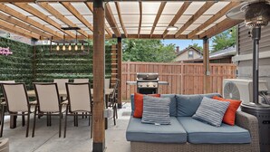 An outdoor patio with a wooden pergola, a blue sofa with cushions, and a dining table with chairs.